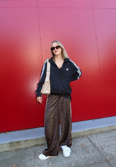 Chaqueta negra con cremallera y rayas blancas, pantalones anchos de estampado de leopardo, y zapatillas blancas. Sostiene un bolso beige y está de pie contra una pared roja.