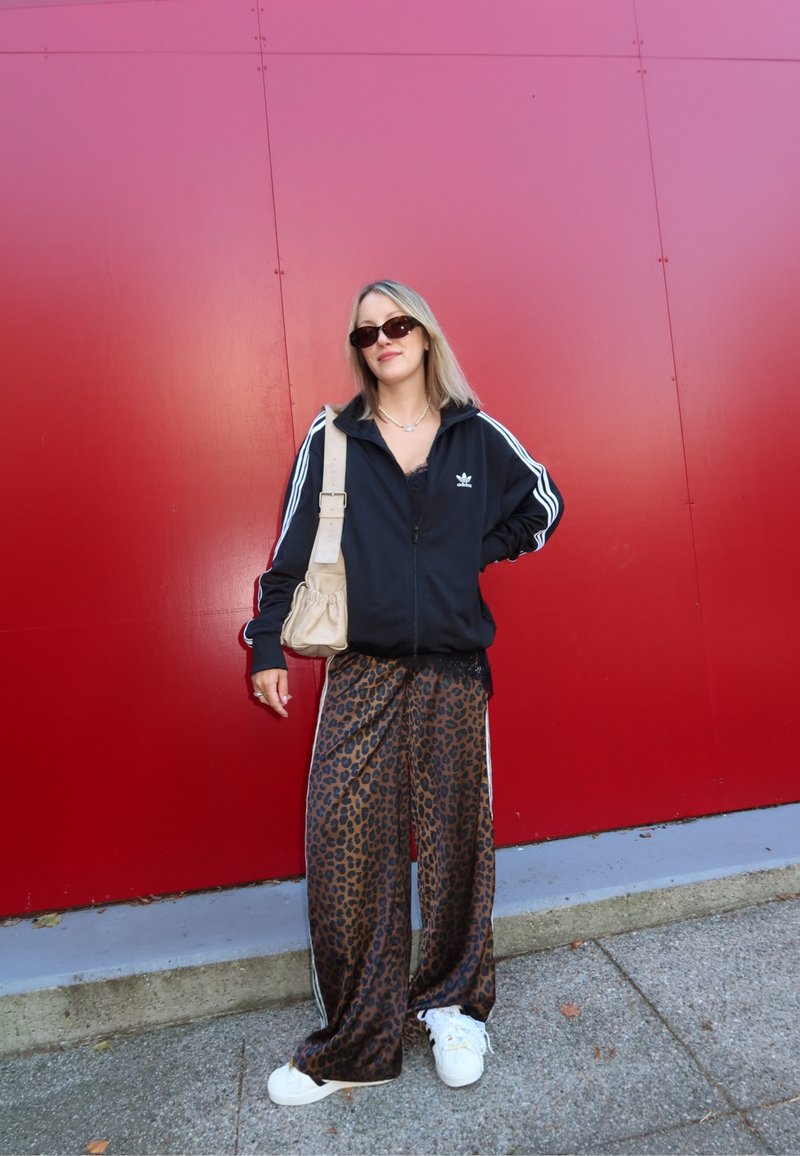 Veste zippée noire avec des rayures blanches, pantalon large en imprimé léopard, et baskets blanches. Tient un sac beige, se tient contre un mur rouge.