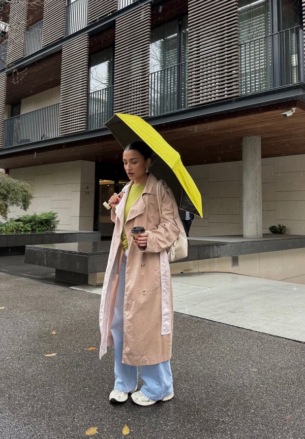 Une femme en manteau beige tient un parapluie jaune vif et un café à emporter sur un trottoir mouillé, devant un bâtiment moderne.