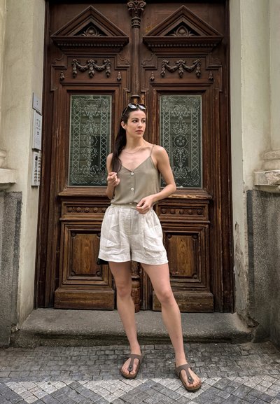 Camiseta de satén en verde oliva claro con botones frontales, combinada con pantalones cortos de lino color blanco roto. La modelo lleva chanclas marrones. Fondo de puerta de madera.