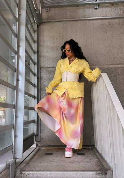 Chaqueta amarilla de satén con botones, cinturilla blanca, falda larga con degradado de rosa a amarillo, zapatillas rosas, de pie en una escalera en un entorno moderno.