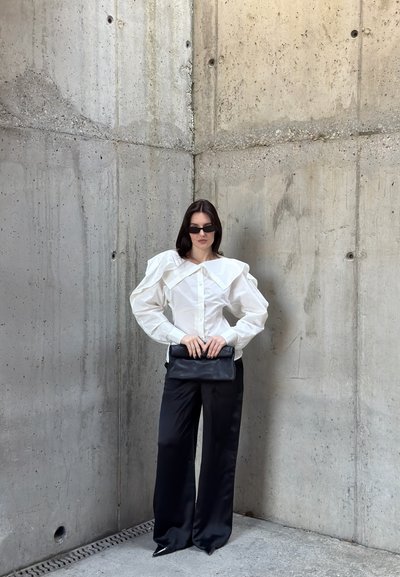 Mujer con cabello oscuro lleva blusa blanca de manga abullonada, pantalones negros de pierna ancha, bolso de mano negro y gafas de sol frente a un fondo de esquina de hormigón.