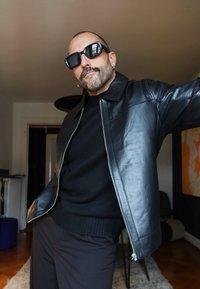 Svart skinnjakke med glidelåslukking, matt tekstur og knappkrage. Brukt over en svart genser. Tilbehør inkluderer solbriller og en øredobb.