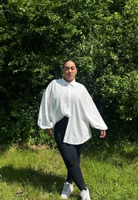 Weißes oversized Hemd mit Knopfverschluss, Kragen und voluminösen Ärmeln, kombiniert mit schwarzen Leggings und weißen Sneakers, vor grünem Hintergrund.