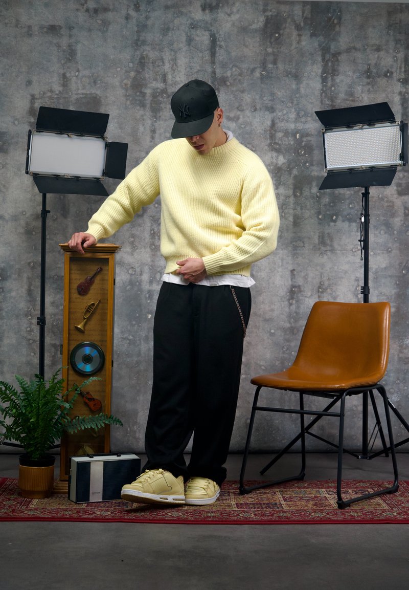 Gelber Rippstrickpullover, schwarze Hose mit Kettendetail und gelbe Sneakers. Im Hintergrund sind eine Holz-Musikanzeige und ein Ledersessel zu sehen.