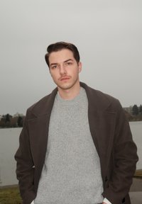 Jeune homme en manteau sombre et pull gris debout près de l'eau par une journée nuageuse, avec des arbres et des bâtiments en arrière-plan.
