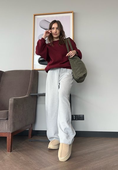 Suéter burdeos, pantalones deportivos grises, calzado beige. Bolsa verde oliva, sillón marrón, obra de arte geométrica en una habitación minimalista. Superficies de madera y textura.