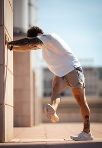 Een persoon in een wit, kort mouw t-shirt en grijze shorts leunt tegen een muur, waarbij een tatoeage op hun been zichtbaar is en ze witte sportschoenen dragen.