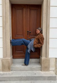 Veste en daim marron, haut noir, écharpe à carreaux et jean évasé en denim. Debout près d'une porte en bois avec une poignée en métal, pied levé sur la marche.