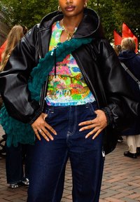 Zwarte leren oversized jas met gathered manchetten, kleurrijke gebloemde top, denimbroek en een gestructureerde teal schoudertas.