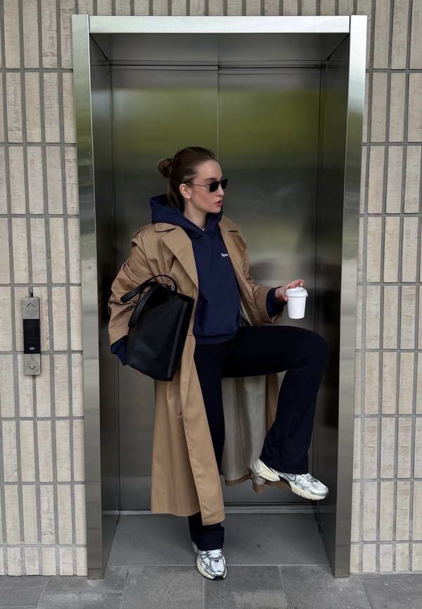 Vrouw draagt een beige trenchcoat, donkerblauwe hoodie, zonnebril en sneakers, houdt een zwarte tas en koffiebeker vast, staat voor een gesloten lift.