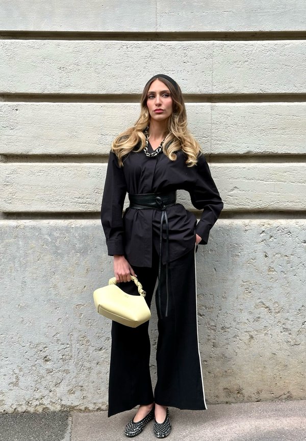 Femme aux longs cheveux ondulés porte une chemise noire ceinturée, un pantalon à jambes larges, des ballerines cloutées, un collier chaîne, et tient un sac à main jaune pâle contre un mur en pierre.