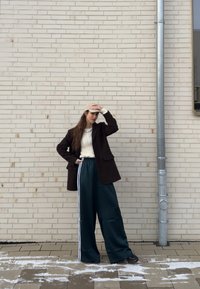 Femme portant une casquette beige, une veste marron foncé, un pull blanc et un pantalon large et foncé, debout contre un mur de briques clair près d'un tuyau argenté.