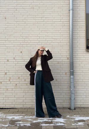 Mujer que lleva una gorra beige, chaqueta marrón oscuro, suéter blanco y pantalones anchos oscuros, de pie junto a una pared clara de ladrillo cerca de una tubería plateada.