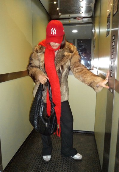 Mujer en un ascensor vestida con un abrigo de piel sintética marrón, una gorra de béisbol roja y una larga bufanda roja, sosteniendo un bolso negro y presionando el botón del ascensor.