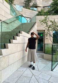 Μαύρη αμάνικη πλεκτή μπλούζα, μπεζ φαρδιές βερμούδες, μαύρα παπούτσια και λευκές κάλτσες. Μοντέρνα πέτρινη αρχιτεκτονική με γυάλινες κιγκλιδώματα και πράσινο.