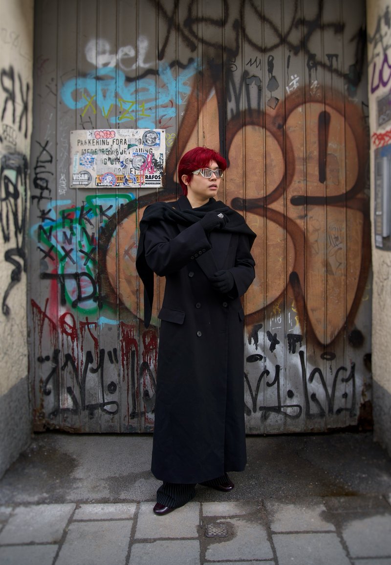 Crni kaput s dvostrukim kopčanjem i ovratnikom, nošen preko crne majice, uparen s hlačama na pruge i sjajnim cipelama. Srebrne sunčane naočale dodaju moderan štih.