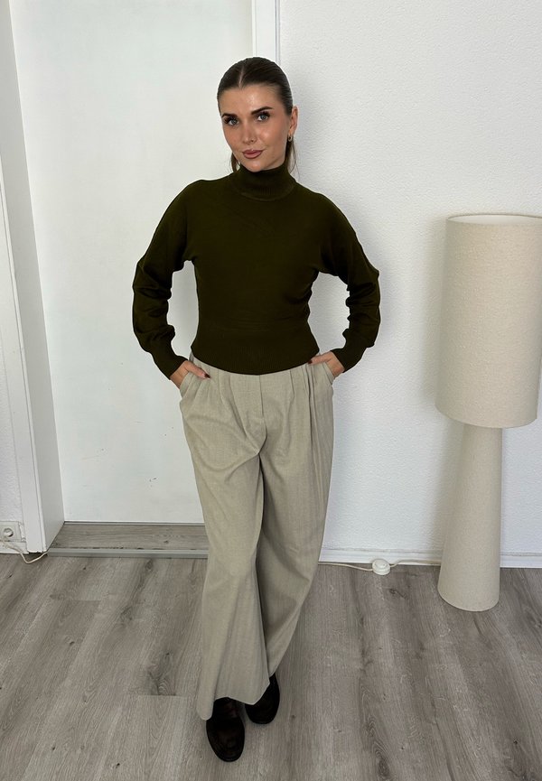 Mujer con cabello oscuro recogido en un moño, vestida con un jersey de cuello alto verde oscuro y pantalones anchos beige, de pie con las manos en los bolsillos dentro de una habitación luminosa con una lámpara.