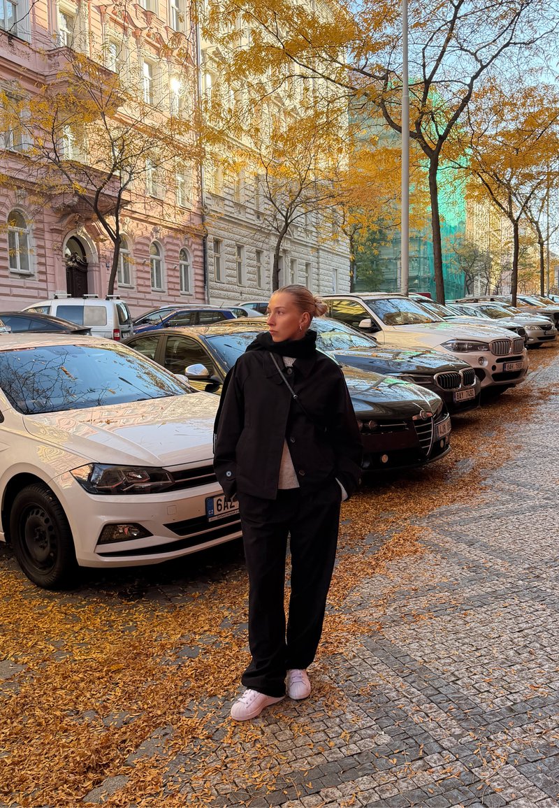 Must musta jope, hele kampsun ja mustad püksid, mida kannab inimene tänaval, kus pargitud autod ja maas sügislehed.