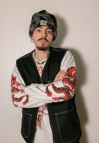 Svart denimväst över vit långärmad skjorta med röd drakgrafik på ärmarna. Mörkmönstrad beanie och färgglad pärlhalsband.