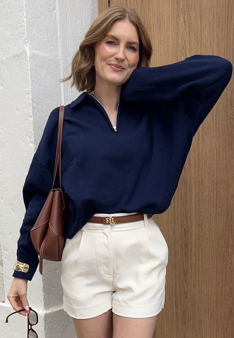 Navy zippered pullover sweater, cream pleated shorts, brown belt with gold hardware, and matching brown handbag. Smooth texture, casual fit.