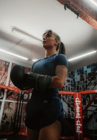 Naine poksija sinises spordiriietuses ja mustade kinnastega seisab jõusaali poksiringis käed rüpes, seinu kaunistavad plakatid.