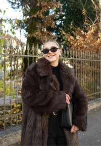 Cappotto in pelliccia sintetica marrone con collo ampio, indossato sopra un dolcevita nero. Gli accessori includono occhiali da sole neri e una piccola borsa nera.