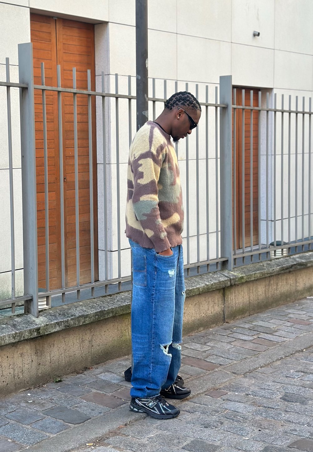 Pull en tricot à motif camouflage beige, marron et vert ; jeans larges bleu clair avec des déchirures ; baskets noires ; lunettes de soleil ; debout à côté d'un mur.