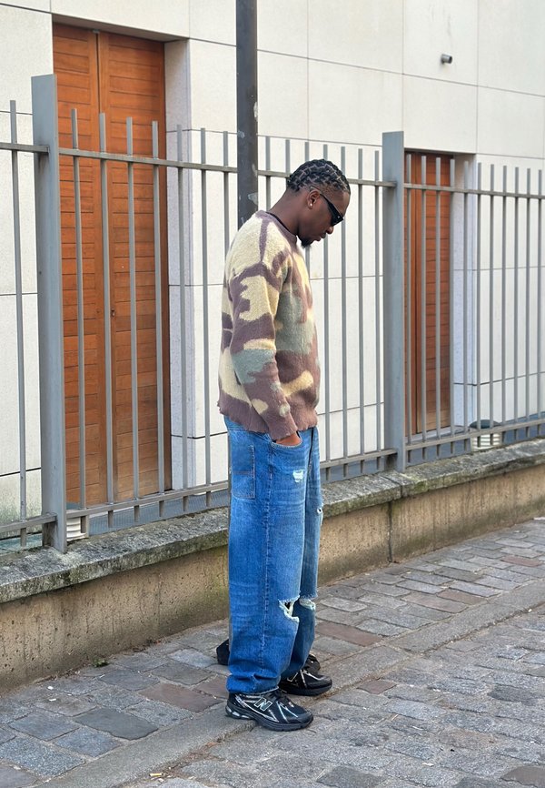 Knitted jumper in beige, brown, and green camouflage pattern; loose-fitting light blue jeans with rips; black trainers; sunglasses; standing beside a wall.