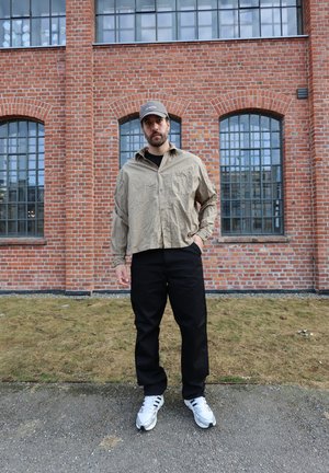 Hombre con camisa de cuadros beige, pantalones negros, gorra gris y zapatillas blancas de pie frente a un edificio de ladrillo con grandes ventanas.