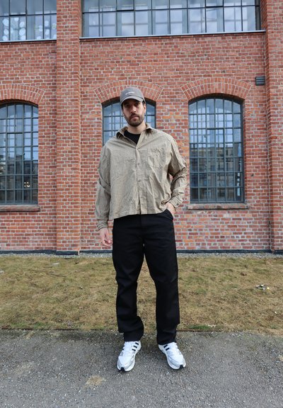 Hombre con camisa de cuadros beige, pantalones negros, gorra gris y zapatillas blancas de pie frente a un edificio de ladrillo con grandes ventanas.