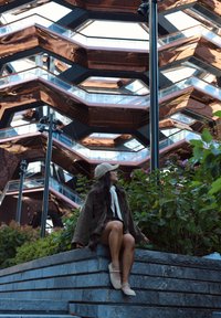 Brun ruskindjakke, grå kasket, blomstergrønt og en tekstureret stensæde. I baggrunden ses en moderne, multifacetteret bygning med glaselementer.