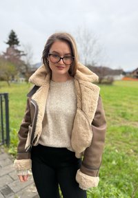 Brun shearling-læderjakke med for af kunstpels, med hætte og kontrastkant, båret over en beige sweater og sorte bukser.
