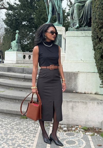 Top negro sin mangas, falda lápiz marrón de cintura alta con abertura frontal, medias negras, tacones negros y un bolso marrón. La joyería añade detalles.