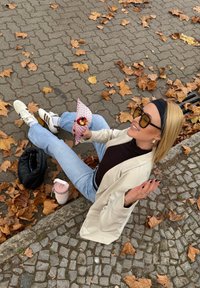 Een lichtbeige blazer over een zwarte coltrui en blauwe spijkerbroek. Witte sneakers met bruine accenten. Houdt een roze hoorntje met fruit vast.