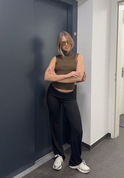 Mujer con blusa marrón sin mangas, pantalones negros, zapatillas blancas y gafas de sol tintadas, de pie con los brazos cruzados junto a una puerta de ascensor oscura.