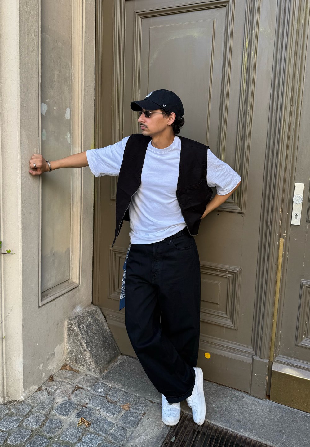 Veste sans manches noire portée sur un t-shirt blanc, associée à un pantalon noir ample et des baskets blanches, accessoirisée avec des lunettes de soleil et une casquette.