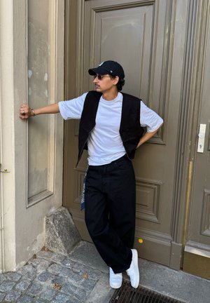 Chaleco negro sin mangas sobre una camiseta blanca, combinado con pantalones negros holgados y zapatillas blancas, accessorios con gafas de sol y una gorra.