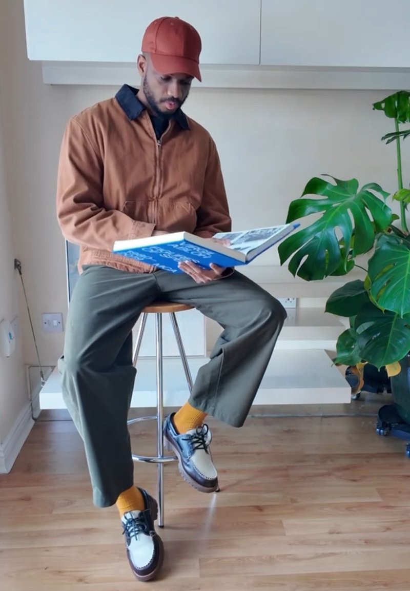 Brown bomber jacket with black collar, olive green pants, bright yellow socks, and brown and white shoes. Reading a blue book while seated.