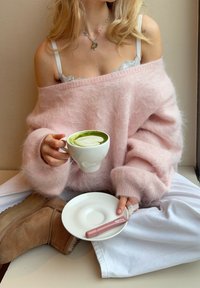 Femme en pull rose pelucheux et pantalon blanc assise en tailleur, tenant une tasse de latte matcha vert et une soucoupe avec un stylo vape rose.