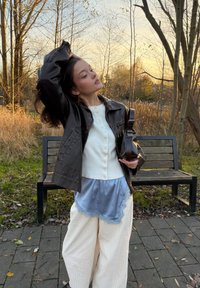 Giacca di pelle nera sopra un maglione bianco con bottoni, stratificata con un top di seta blu. Pantaloni larghi color crema e borsa scura in mano.