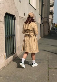 Manteau beige avec une taille ceinturée, associé à des chaussettes blanches à motifs et des baskets blanches avec des accents verts, sur une rue pavée.