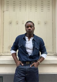Homme aux cheveux tressés portant une chemise en jean bleu par-dessus un henley blanc, un jean foncé et des accessoires, se tenant contre un mur beige avec des volets.