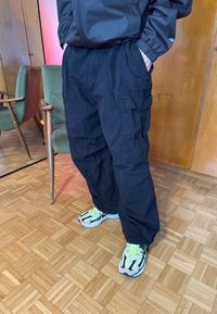 Personne portant un pantalon cargo noir, une veste noire avec les mains dans les poches et des baskets blanches, debout sur un sol en parquet près de chaises vertes et rouges.