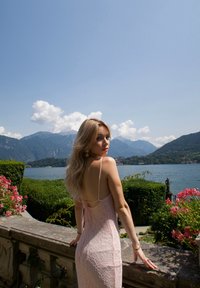 Robe en dentelle rose clair avec un dos décolleté et de fines bretelles, mise en scène au bord d'un lac entouré de montagnes et de fleurs en fleurs.