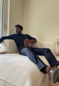 Mann in dunklem Pullover und Hose, der auf einem weißen Sofa liegt, eine braune Handtasche hält und in einem hellen Raum mit strukturierten Wänden aus dem Fenster schaut.