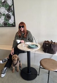 Mesa redonda com tampo de mármore apoiada numa base preta, uma mala de couro castanho, um prato verde-claro, um copo e um cão pequeno no chão.
