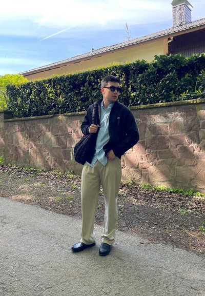 Persona vestida con una chaqueta negra de cremallera, camisa de botones azul claro, pantalones beige y zapatos oscuros, sosteniendo una bolsa de hombro acolchada al aire libre.