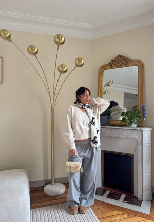 Suéter grueso de punto blanco, pantalones grises, bufanda floral en blanco y negro, bolso beige, lámpara de pie dorada, chimenea de mármol, suelo de madera.