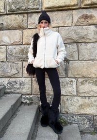 Giacca in pile color crema con collo alto, berretto nero, leggings neri e stivali neri rifiniti con pelliccia, in piedi contro un muro di pietra.